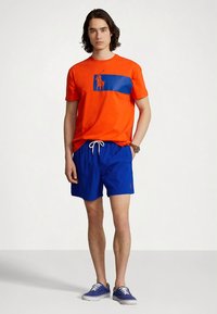 Młody mężczyzna w jasnopomarańczowej koszulce z niebieskim logo, niebieskich spodenkach z troczkami i niebieskich sneakersach, stojący na tle białym.