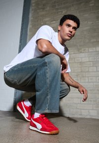 Red suede sneakers with white accents and gum sole, worn with blue denim jeans and a white t-shirt, against a textured brick wall.