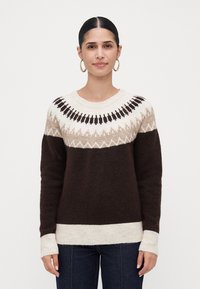 Kvinne med mørkt hår i en lav knute, iført gull hoop-øredobber og en strikkegenser med brune, beige og hvite mønstre, samt mørke jeans.