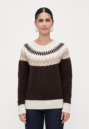 Kvinne med mørkt hår i en lav knute, iført gull hoop-øredobber og en strikkegenser med brune, beige og hvite mønstre, samt mørke jeans.