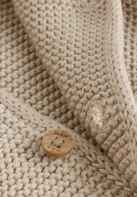 Pull en maille beige avec un motif texturé, agrémenté d'un bouton en bois gravé "NEXT". Le tissu présente une apparence douce et tissée.