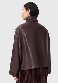 Donna con capelli neri corti che indossa una giacca in pelle marrone scuro e una gonna plissè, vista di spalle su uno sfondo chiaro.