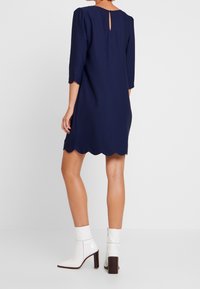 Marinblå klänning med trekvartslånga puffärmar, vågig fåll och nyckelhålsdetalj bak. Har en slät textur och bärs med vita ankelskor.