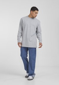 Camiseta de manga larga gris claro, de corte relajado, con cuello redondo. Combinada con unos pantalones vaqueros azules holgados y zapatillas blancas en un entorno plano y neutral.