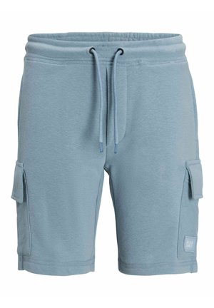Hvid T-shirt med logo, lyseblå shorts med lommer og beige sneakers. Sorte sokker synlige, kort krøllet hår. Neutral baggrund.