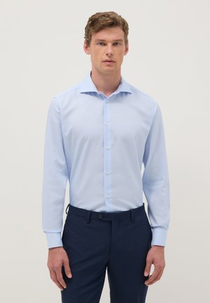 Man draagt een lichtblauw overhemd met knopen, ingestopt in donkerblauwe broek, staand tegen een effen lichte achtergrond.