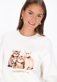 Fehér pulóver három masnis kiscicával, nyomtatott grafika, kerek nyakkivágás és bordázott mandzsetták. Alatta a felirat: „Erősen tartom magam.”