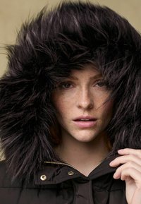 Schwarze Pufferjacke mit einer großen, flauschigen schwarzen Kunstfellkapuze. Der Reißverschluss ist goldfarben und der Stoff wirkt glatt mit einem leichten Glanz.