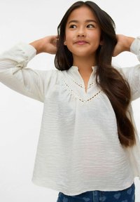 Blusa bianca a maniche lunghe con scollo a V e dettagli in pizzo intricati. Tessuto morbido e strutturato con vestibilità ampia. Abbinata a jeans scuri.