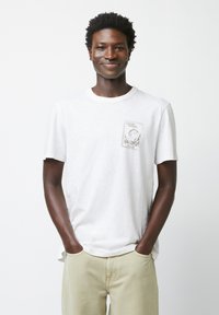 Weißes T-Shirt mit kurzen Ärmeln aus leichtem Stoff, mit einem Fronttaschen-Design und grüner Grafik, die "Fishing Therapy" sagt.