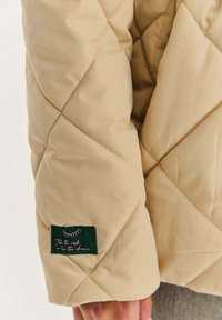 Rukáv béžové prošívané bundy se zelenou nášivkou s růžovým textem. Hladká textura, vzor ve tvaru diamantů a viditelný detail šití.