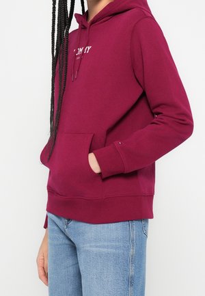 Personne portant un sweat à capuche bordeaux avec poche avant et un jean bleu clair, main dans la poche, cheveux tressés tombant sur l’épaule.