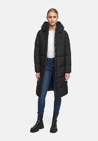 Sort pufferjakke med hætte, lang længde, quiltet design, foran lynlåslukning, båret over en hvid top og blå jeans, parret med sorte støvler.