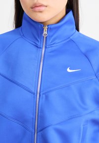 Blå syntetisk jakke med glidelås og høy krage, med hvit Nike-logo på venstre bryst og teksturert detailing på stoffet.