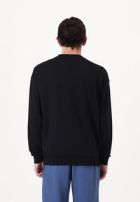 Sudadera negra con cuello redondo, puños y bajo de canalé. Textura suave, corte relajado, vista desde la parte de atrás, combinada con pantalones de color azul claro.