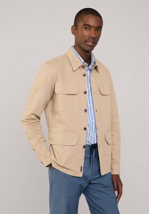 Beige katoenen jack met een kraag, sluiting met knopen aan de voorkant, twee borstzakken en een slim fit. Draag het over een blauw-wit gestreept shirt.