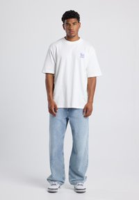 White cotton t-shirt with short sleeves, featuring a small blue graphic on the left chest. Paired with light blue denim jeans and black sneakers.
