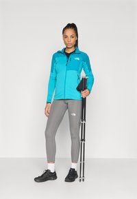 Τουρκουάζ φλις μπουφάν με υφασμάτινο σχέδιο, γκρι κολάν και μαύρα παπούτσια πεζοπορίας. Το μοντέλο κρατά ράβδους πεζοπορίας, στέκεται μπροστά σε ουδέτερο φόντο.