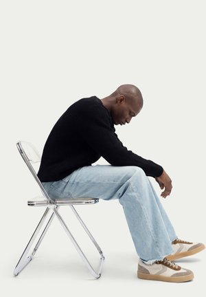 Homme portant un pull noir, un jean bleu clair et des baskets beiges, assis sur une chaise pliante transparente, penché en avant, la tête baissée.
