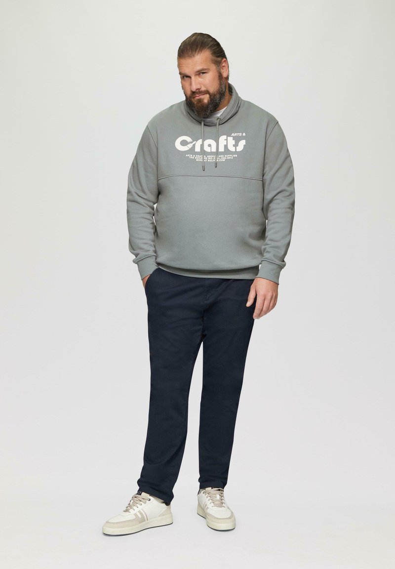 Szara bluza z białym logo, ciemne spodnie i kremowe sneakersy. Bluza ma wysoki kołnierz i sznurki do regulacji. Gładka faktura, casualowy design.