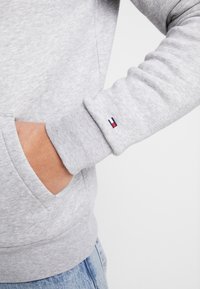 Sudadera gris con un tejido texturizado, que presenta un bolsillo frontal y un pequeño detalle de logo en rojo, blanco y azul en el puño.