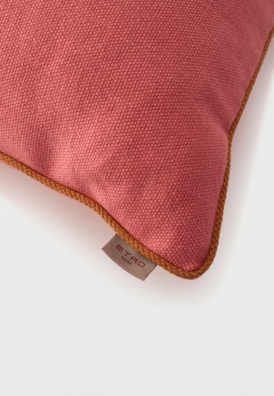Strukturiertes staubrosafarbenes Kissen mit braun geflochtenem Rand und einem kleinen Stoffetikett mit der Aufschrift „ETRO HOME“ auf weißem Hintergrund.