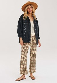 Žena s bež širokým kloboukem, černou oversized bundou, bílým topem, vzorovanými širokými kalhotami a hnědými žabkami, stojící na bílém pozadí.