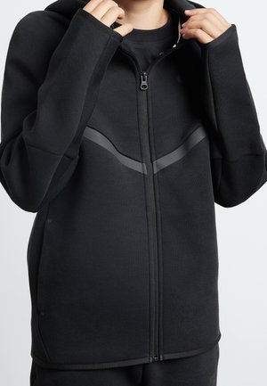 Zwarte hoodie met rits en een gladde textuur, voorzien van een gebogen grijze streep op de borst, elastische manchetten en een ruime capuchon.