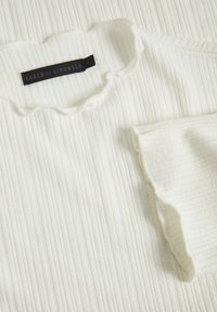 Top in tessuto bianco a costine con bordi arricciati. Presenta un'etichetta nera con la scritta "KAREN by SIMONSEN" e taglia 'M'. Dettaglio della superficie testurizzata.