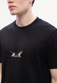 Svart bomull T-skjorte med rund hals, med en brodert design av to fugler og ordet "HUGO" i midten.