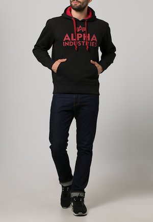 Schwarzer Hoodie mit roten Akzenten und "ALPHA INDUSTRIES" Logo. Rippbündchen und Saum, Kängurutasche, kombiniert mit dunklen Jeans und Sneakers.