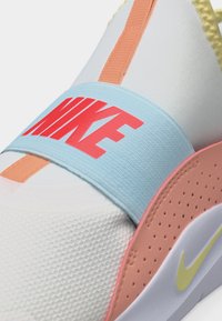 Κοντινό πλάνο σε λευκό αθλητικό παπούτσι με ανοιχτό μπλε λουρί που φέρει κόκκινο λογότυπο Nike, ροδακινί λεπτομέρειες και μικρό κίτρινο σήμα Nike στη σόλα.