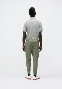 Mees seisab seljaga vaataja poole, kandes halli polo särk, oliivirohelisi lahtisi pükse elastsete mansettidega, musti sokke ja valgeid tossusid.