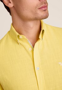Geel overhemd met knopen, gemaakt van textiel met structuur; heeft een contrasterende patroon aan de binnenkant van de kraag en twee blauwe knopen. Subtiel logo op de borst.