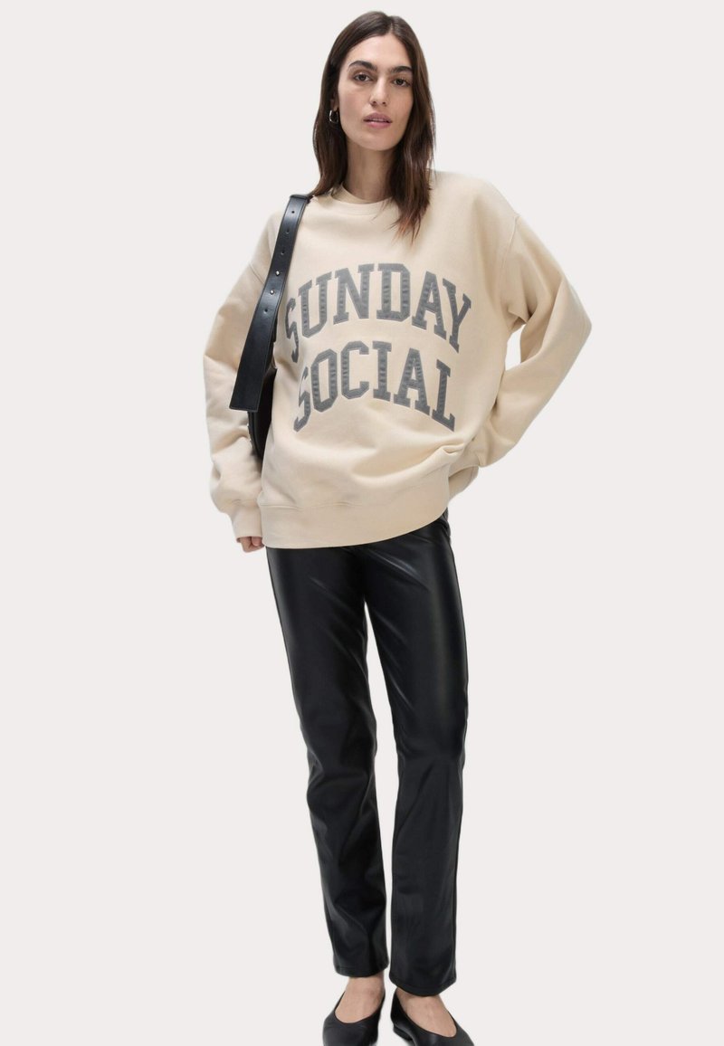 Sweatshirt beige con la scritta "SUNDAY SOCIAL" stampata in grigio. Abbinato a pantaloni in pelle sintetica nera. Tessuto morbido, vestibilità rilassata e maniche lunghe.