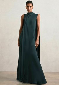 Langes, ärmelloses Kleid aus tiefgrünem Stoff mit einem drapierten Design, hohem Halsausschnitt und einer offenen, kapuzenartigen Silhouette, die von den Schultern ausgeht.