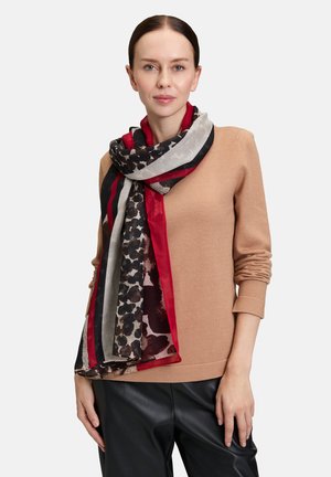 Frau trägt einen braunen Pullover und schwarze Hosen, mit einem rot-schwarz-grauem und leopardgemustertem Schal um den Hals gewickelt.