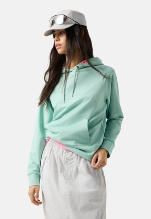 Vrouw draagt een lichtmintgroene hoodie, witte rok met ritszakken, witte baseballpet en zonnebril die op de pet rust, kijkt opzij.