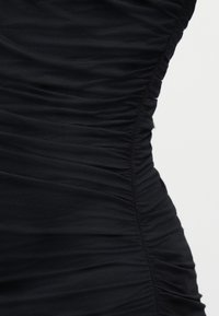 Κοντινό πλάνο μαύρου υφάσματος με ρυτιδώσεις που δημιουργούν συσσωρευμένη υφή και λεπτή σιλουέτα στη ραφή του ενδύματος στο πλάι.