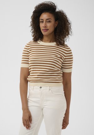 Vrouw met krullend haar die een kort mouwig bruin-wit gestreept shirt draagt en een witte broek met hoge taille, tegen een effen achtergrond.