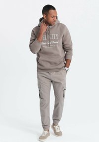 Hoodie i taupe med vit text, kombinerad med ljusgrå cargobyxor med sidofickor och svarta dragkedjor, avslutad med ljusa sneakers.