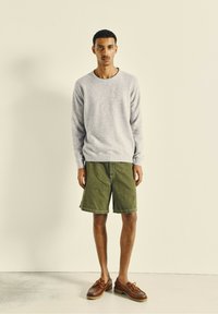 Un pull gris au toucher doux, un short vert olive et des chaussures bateau en cuir marron. La tenue est simple et décontractée, avec une silhouette classique.