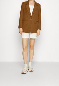 Brun kavaj med en knapp, två framfickor, tillsammans med vita shorts och krämfärgade snörskor, stående på en neutral bakgrund.