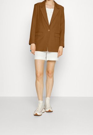 Blazer marron à un bouton, deux poches avant, associé à un short blanc et des baskets crème à lacets, se tenant sur un fond neutre.
