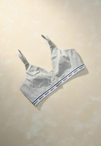 Sivý bavlnený bralet s tenkými ramienkami, s širokým elastickým pásom s textom "NEXT" v čiernej farbe na bielom pozadí. Hladká textúra.