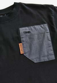 Sort t-shirt med en grå brystlomme med lynlås, akkompagneret af et orange "SUBSCRIBE"-mærke. 100% bomuldsstof, ribbet halsudskæring.