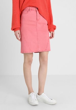 Femme portant une jupe rose longueur genou, un pull en tricot orange et des baskets blanches, debout devant un fond clair uni.