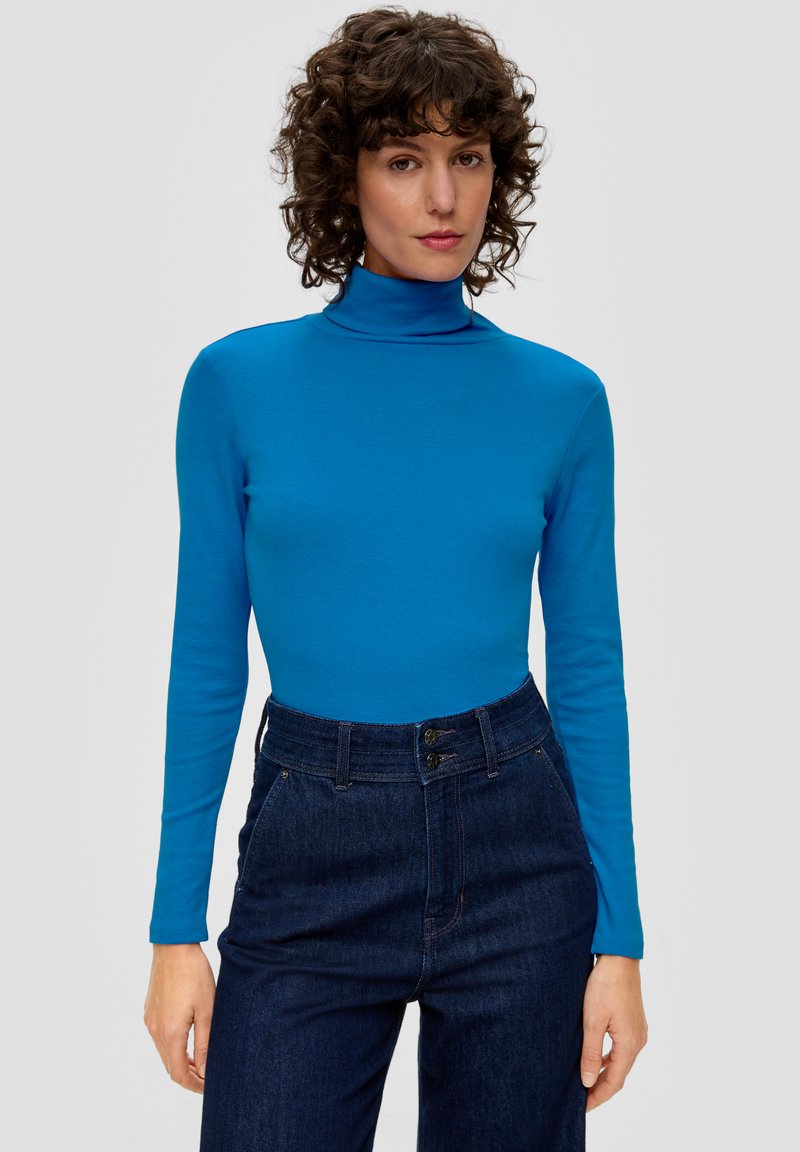 Blauwe turtleneck met lange mouwen van soepel materiaal, gecombineerd met donkere high-waisted denim broek met sluiting met twee knopen.