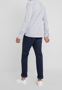 Jasnoszara koszula z długim rękawem z kołnierzykiem zapinanym na guziki, zestawiona z granatowymi spodniami i białymi trampkami. Posiada podwinięte mankiety i gładką fakturę.