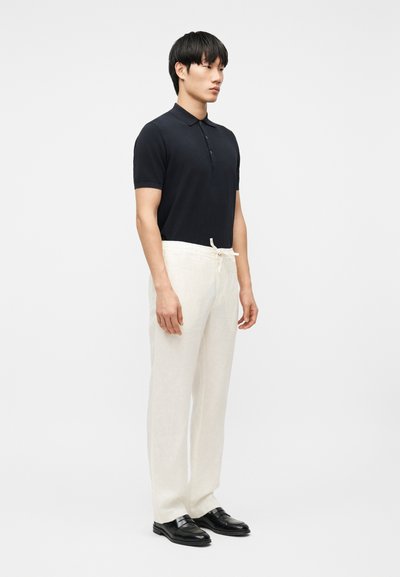 Άνδρας που στέκεται φορώντας μαύρο μπλουζάκι πόλο με κοντά μανίκια, λευκό παντελόνι με κορδόνι και μαύρα επίσημα παπούτσια μπροστά σε απλό φωτεινό φόντο.